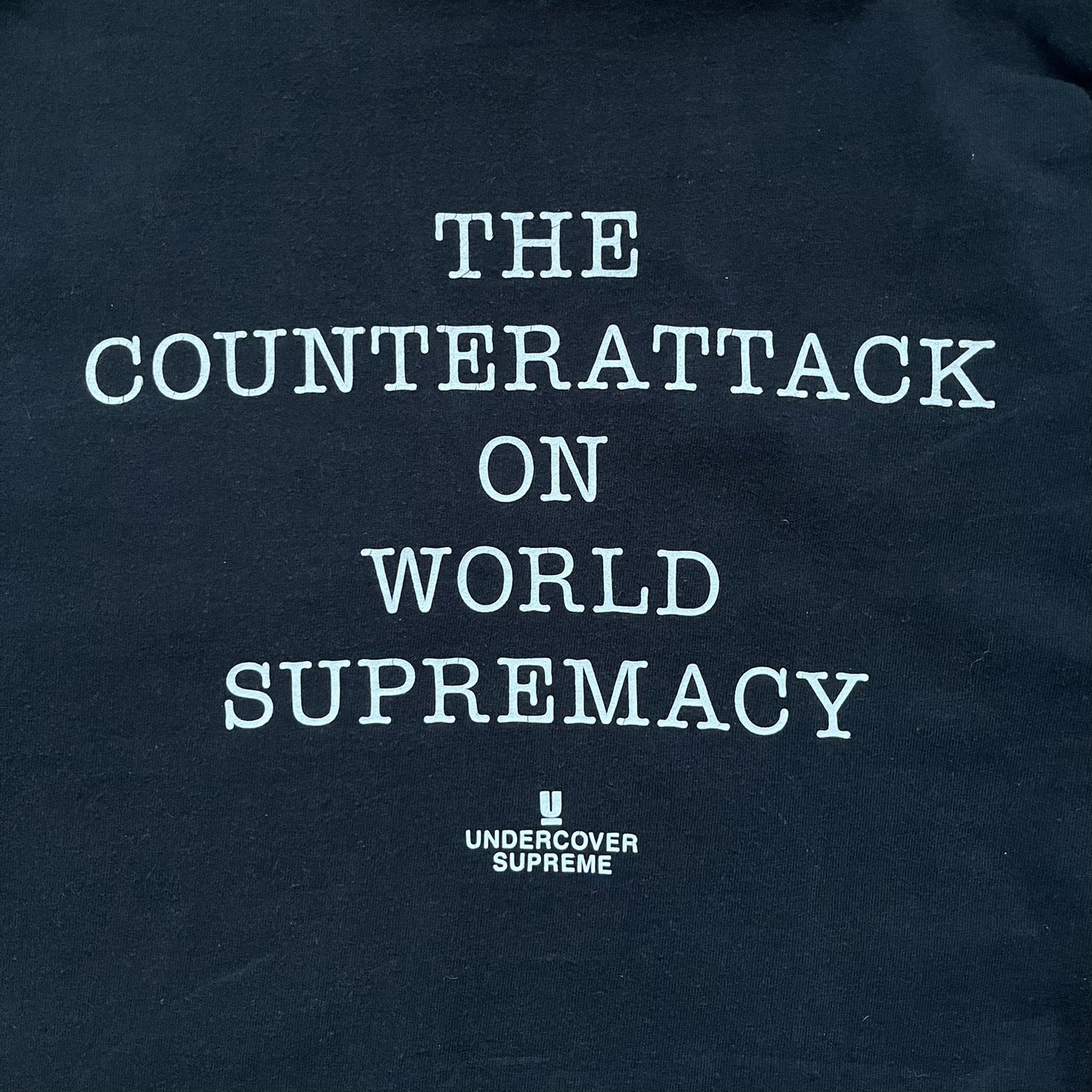 Supreme Undercover Public Enemy Counterattack SS18 Longsleeve Tee is a black long-sleeve T-shirt released on March 15, 2018, crafted from 100% cotton. The front features white “The Counterattack On World Supremacy” text with a small Supreme UNDERCOVER logo beneath, while the back displays a large Public Enemy “Fear of a Black Planet” graphic with galaxy and planet imagery and the same co-branded logo below. The design includes a ribbed crewneck and ribbed cuffs, reflecting the SS18 collaboration between Sup