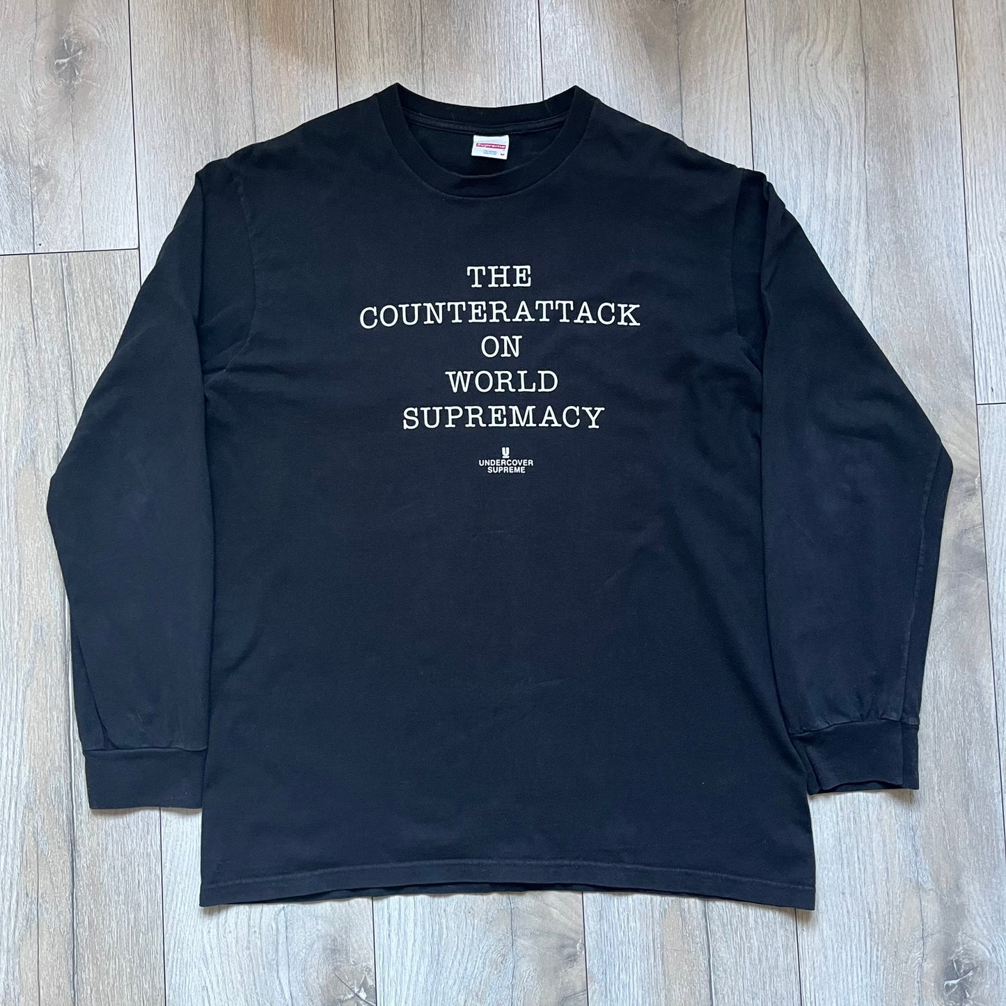 Supreme Undercover Public Enemy Counterattack SS18 Longsleeve Tee is a black long-sleeve T-shirt released on March 15, 2018, crafted from 100% cotton. The front features white “The Counterattack On World Supremacy” text with a small Supreme UNDERCOVER logo beneath, while the back displays a large Public Enemy “Fear of a Black Planet” graphic with galaxy and planet imagery and the same co-branded logo below. The design includes a ribbed crewneck and ribbed cuffs, reflecting the SS18 collaboration between Sup