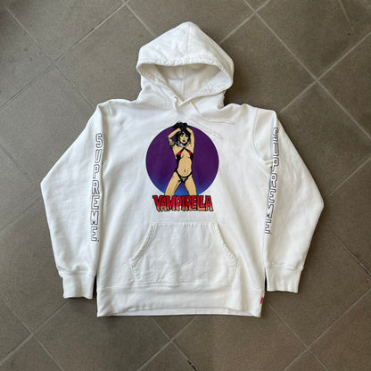 Supreme Vampirella SS17 White Hoodie features a white 100% cotton construction with a front graphic of Vampirella against a purple circular background. A “Vampirella” logo is printed underneath, while black outlined Supreme logos run vertically along the sleeves.

