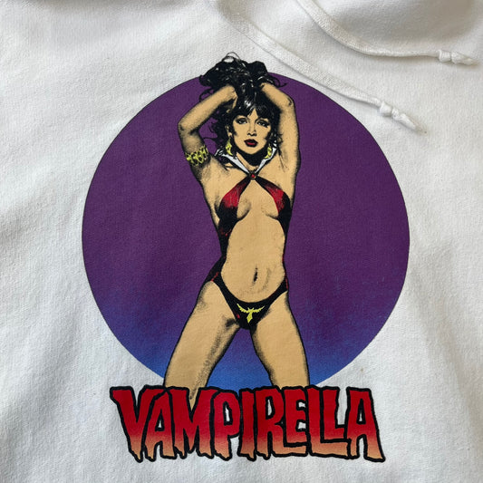 Supreme Vampirella SS17 White Hoodie features a white 100% cotton construction with a front graphic of Vampirella against a purple circular background. A “Vampirella” logo is printed underneath, while black outlined Supreme logos run vertically along the sleeves.
