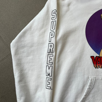 Supreme Vampirella SS17 White Hoodie features a white 100% cotton construction with a front graphic of Vampirella against a purple circular background. A “Vampirella” logo is printed underneath, while black outlined Supreme logos run vertically along the sleeves.
