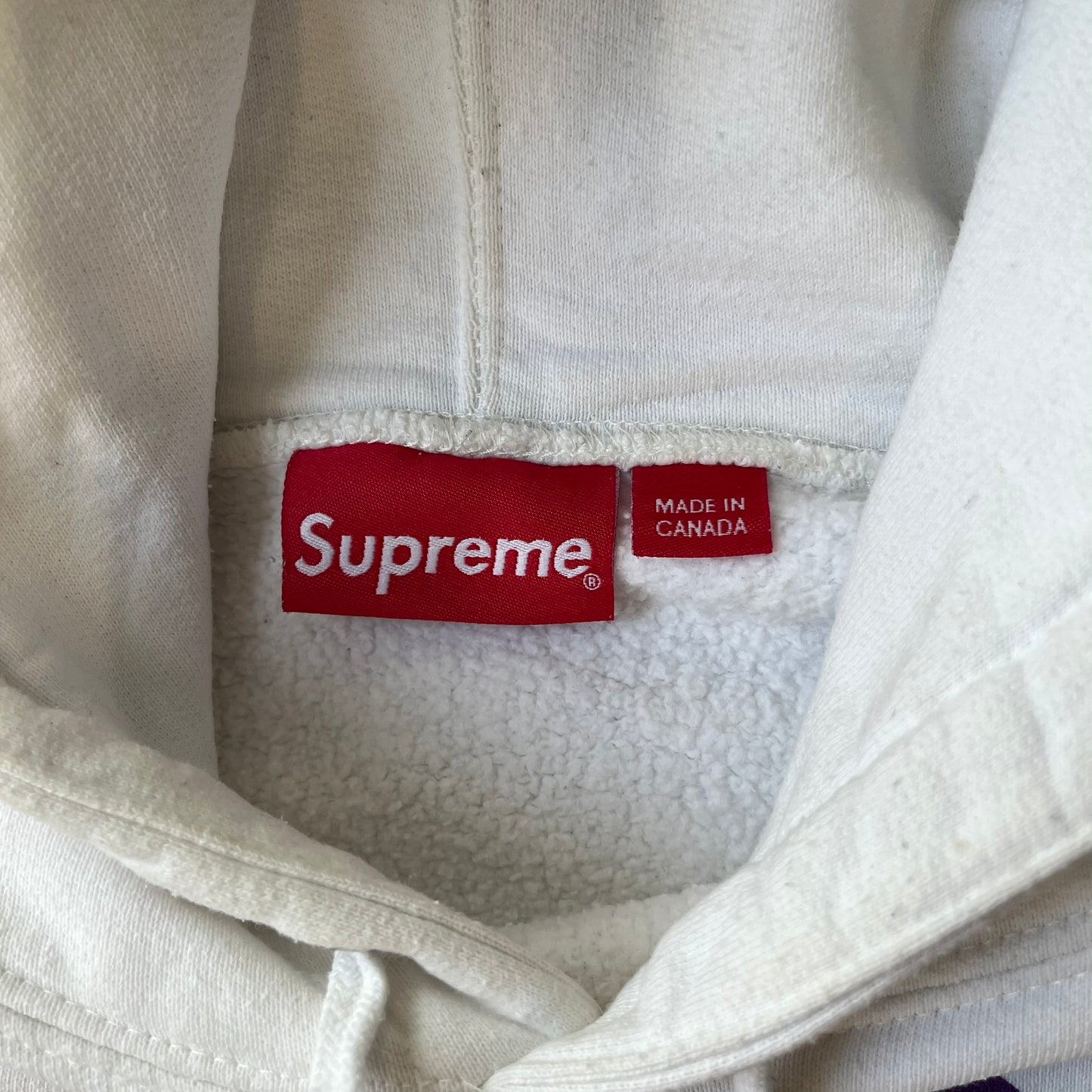 Supreme Vampirella SS17 White Hoodie features a white 100% cotton construction with a front graphic of Vampirella against a purple circular background. A “Vampirella” logo is printed underneath, while black outlined Supreme logos run vertically along the sleeves.
