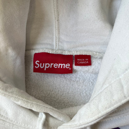 Supreme Vampirella SS17 White Hoodie features a white 100% cotton construction with a front graphic of Vampirella against a purple circular background. A “Vampirella” logo is printed underneath, while black outlined Supreme logos run vertically along the sleeves.
