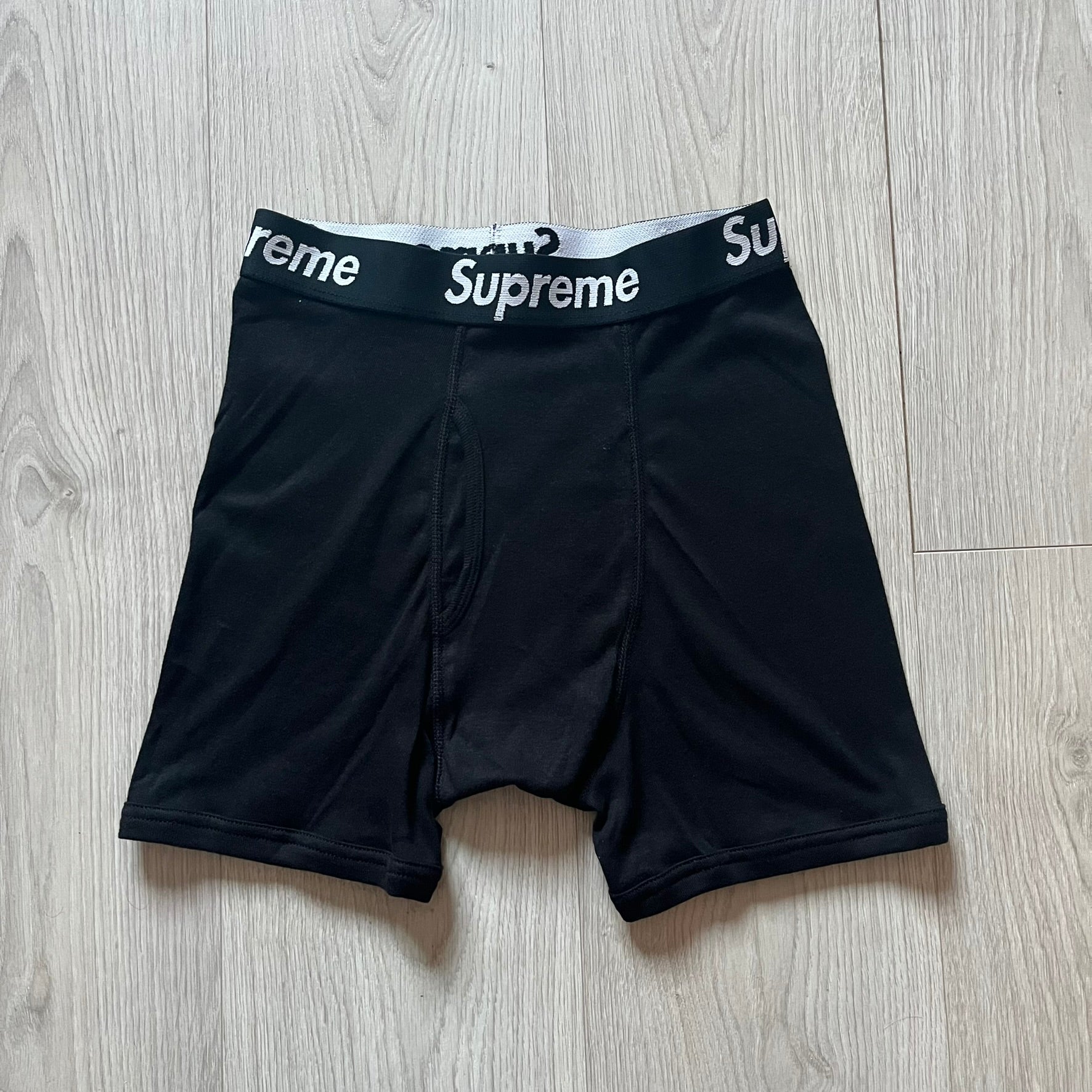 Supreme x Hanes black boxer briefs made of cotton with a stretch fit. Features a black elastic waistband with white Supreme logo text in Futura Oblique font. Classic Hanes base model with co-branded detailing.