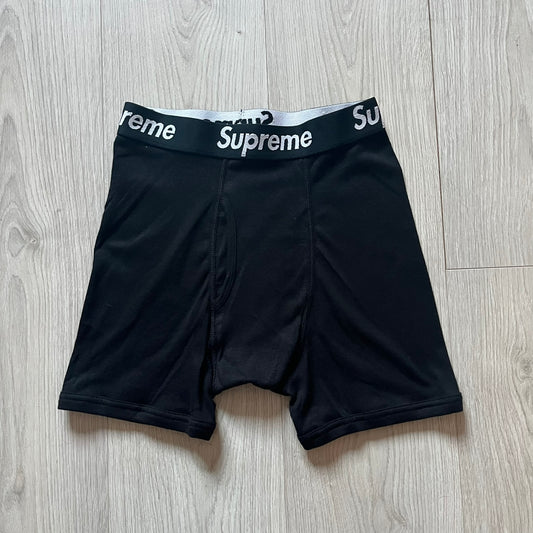 Supreme x Hanes black boxer briefs made of cotton with a stretch fit. Features a black elastic waistband with white Supreme logo text in Futura Oblique font. Classic Hanes base model with co-branded detailing.