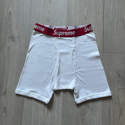 Supreme x Hanes white boxer briefs made of cotton with a stretch fit. Features a red elastic waistband with white Supreme logo text in Futura Oblique font. Classic Hanes base model with co-branded detailing.