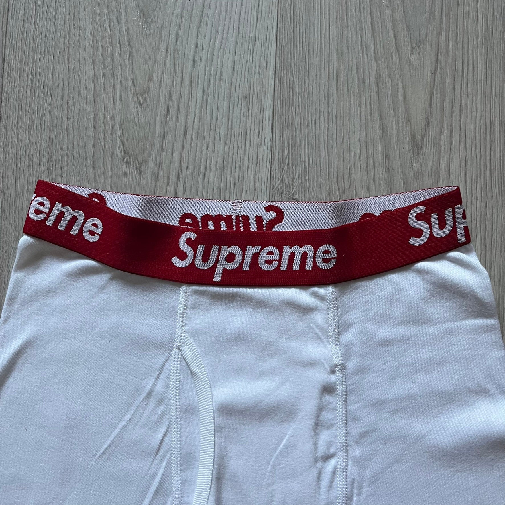 Supreme x Hanes white boxer briefs made of cotton with a stretch fit. Features a red elastic waistband with white Supreme logo text in Futura Oblique font. Classic Hanes base model with co-branded detailing.
