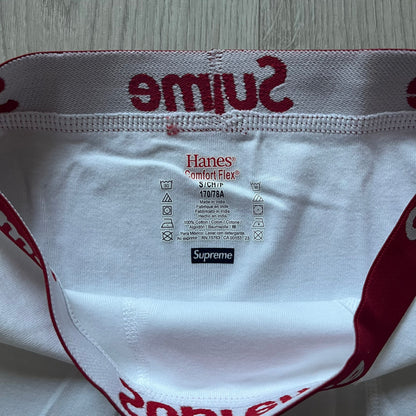 Supreme x Hanes white boxer briefs made of cotton with a stretch fit. Features a red elastic waistband with white Supreme logo text in Futura Oblique font. Classic Hanes base model with co-branded detailing.
