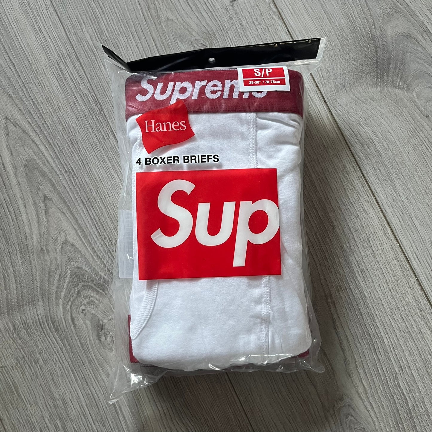Supreme x Hanes white boxer briefs made of cotton with a stretch fit. Features a red elastic waistband with white Supreme logo text in Futura Oblique font. Classic Hanes base model with co-branded detailing.