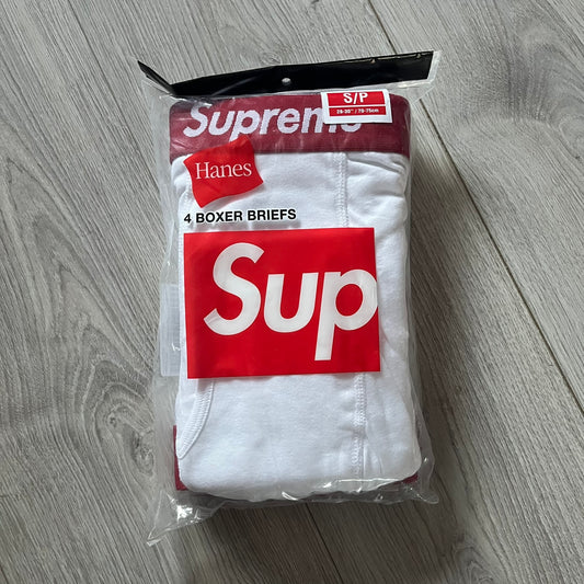 Supreme x Hanes white boxer briefs made of cotton with a stretch fit. Features a red elastic waistband with white Supreme logo text in Futura Oblique font. Classic Hanes base model with co-branded detailing.