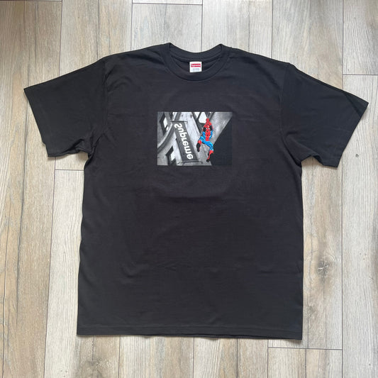 Supreme x Spider-Man Black Tee from the SS26 season is a black 100% cotton T-shirt featuring a large box graphic on the front. The print shows a black and white city photo with the Supreme store, while Spider-Man in his red and blue costume swings across the scene in a cartoon-style illustration. The shirt includes a Supreme x Marvel collaboration neck tag.&nbsp;