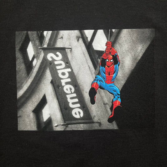 Supreme x Spider-Man Black Tee from the SS26 season is a black 100% cotton T-shirt featuring a large box graphic on the front. The print shows a black and white city photo with the Supreme store, while Spider-Man in his red and blue costume swings across the scene in a cartoon-style illustration. The shirt includes a Supreme x Marvel collaboration neck tag.&nbsp;