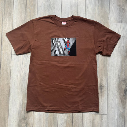 Supreme x Spider-Man Black Tee from the SS26 season is a brown 100% cotton T-shirt featuring a large box graphic on the front. The print shows a black and white city photo with the Supreme store, while Spider-Man in his red and blue costume swings across the scene in a cartoon-style illustration. The shirt includes a Supreme x Marvel collaboration neck tag.