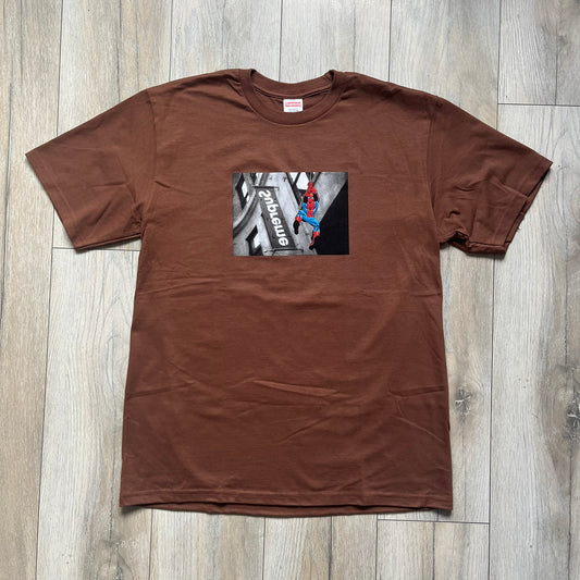 Supreme x Spider-Man Black Tee from the SS26 season is a brown 100% cotton T-shirt featuring a large box graphic on the front. The print shows a black and white city photo with the Supreme store, while Spider-Man in his red and blue costume swings across the scene in a cartoon-style illustration. The shirt includes a Supreme x Marvel collaboration neck tag.