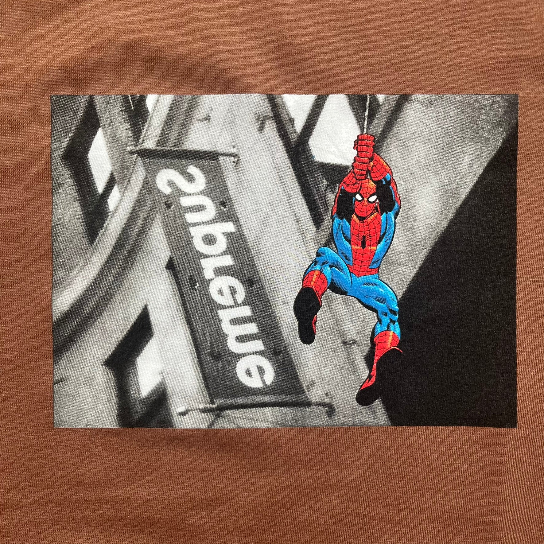 Supreme x Spider-Man Black Tee from the SS26 season is a brown 100% cotton T-shirt featuring a large box graphic on the front. The print shows a black and white city photo with the Supreme store, while Spider-Man in his red and blue costume swings across the scene in a cartoon-style illustration. The shirt includes a Supreme x Marvel collaboration neck tag.
