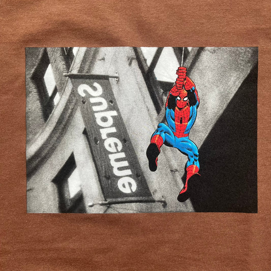 Supreme x Spider-Man Black Tee from the SS26 season is a brown 100% cotton T-shirt featuring a large box graphic on the front. The print shows a black and white city photo with the Supreme store, while Spider-Man in his red and blue costume swings across the scene in a cartoon-style illustration. The shirt includes a Supreme x Marvel collaboration neck tag.