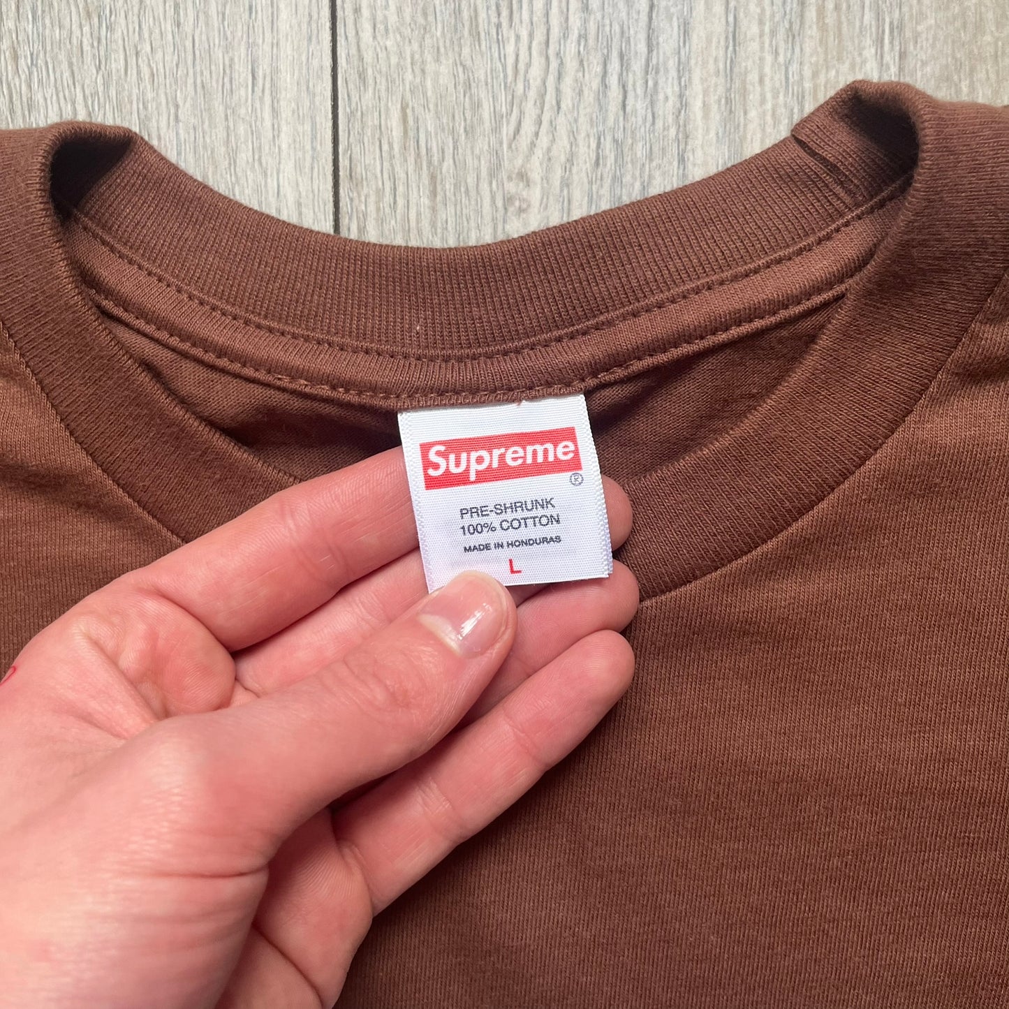 Supreme x Spider-Man Black Tee from the SS26 season is a brown 100% cotton T-shirt featuring a large box graphic on the front. The print shows a black and white city photo with the Supreme store, while Spider-Man in his red and blue costume swings across the scene in a cartoon-style illustration. The shirt includes a Supreme x Marvel collaboration neck tag.