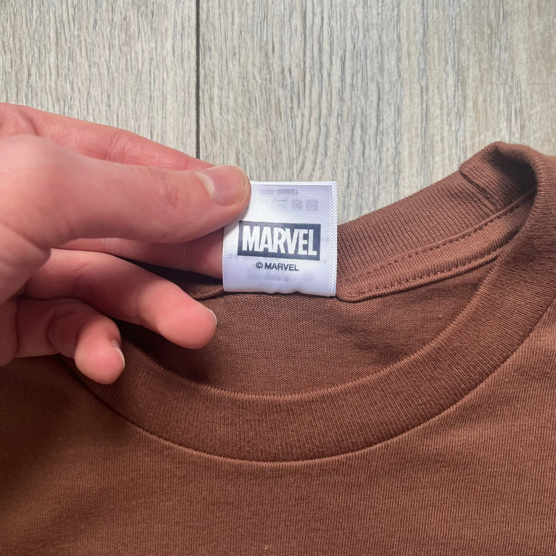 Supreme x Spider-Man Black Tee from the SS26 season is a brown 100% cotton T-shirt featuring a large box graphic on the front. The print shows a black and white city photo with the Supreme store, while Spider-Man in his red and blue costume swings across the scene in a cartoon-style illustration. The shirt includes a Supreme x Marvel collaboration neck tag.