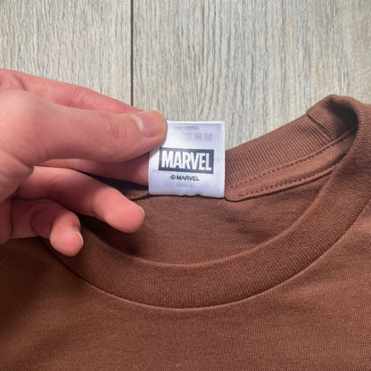 Supreme x Spider-Man Black Tee from the SS26 season is a brown 100% cotton T-shirt featuring a large box graphic on the front. The print shows a black and white city photo with the Supreme store, while Spider-Man in his red and blue costume swings across the scene in a cartoon-style illustration. The shirt includes a Supreme x Marvel collaboration neck tag.