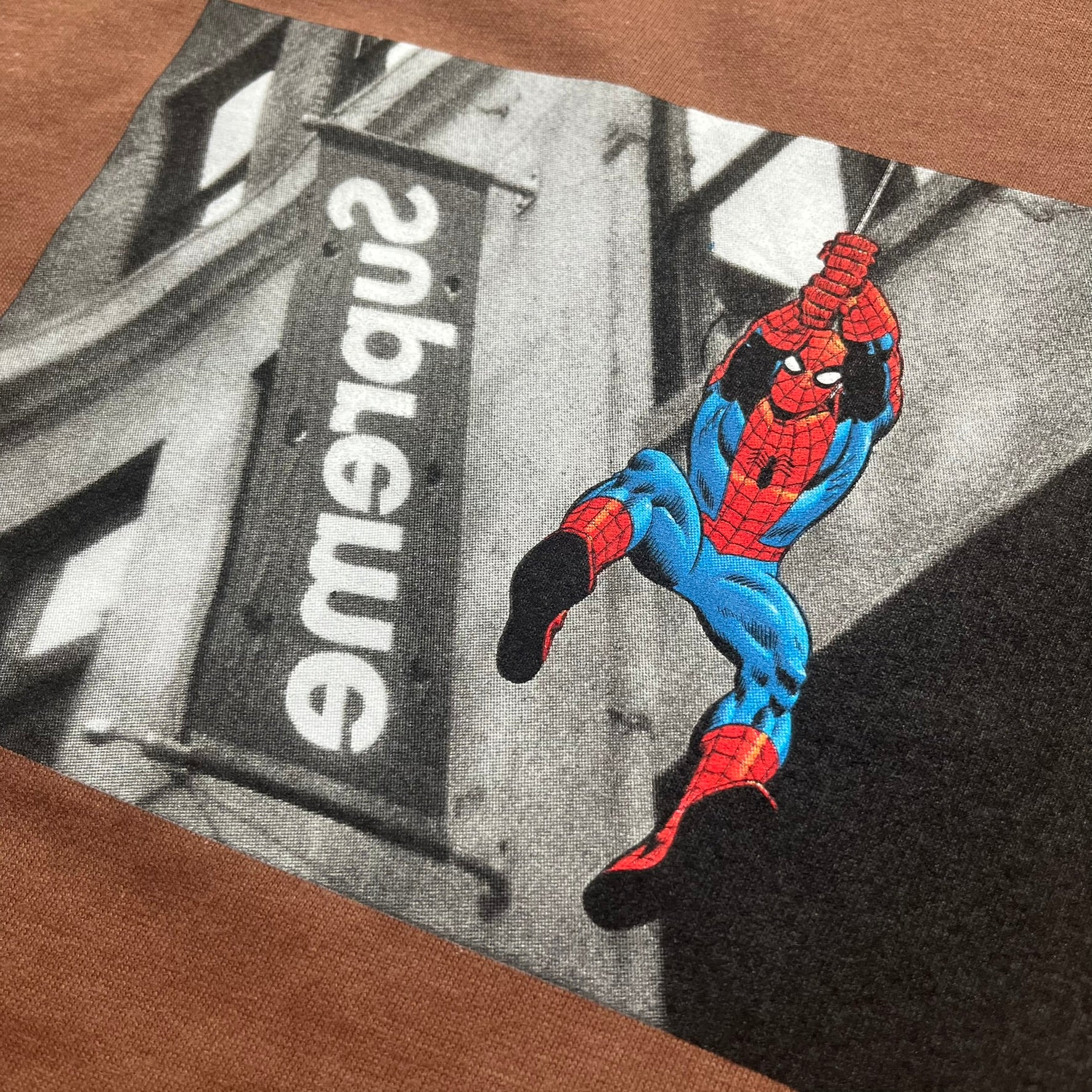 Supreme x Spider-Man Black Tee from the SS26 season is a brown 100% cotton T-shirt featuring a large box graphic on the front. The print shows a black and white city photo with the Supreme store, while Spider-Man in his red and blue costume swings across the scene in a cartoon-style illustration. The shirt includes a Supreme x Marvel collaboration neck tag.