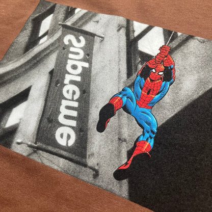 Supreme x Spider-Man Black Tee from the SS26 season is a brown 100% cotton T-shirt featuring a large box graphic on the front. The print shows a black and white city photo with the Supreme store, while Spider-Man in his red and blue costume swings across the scene in a cartoon-style illustration. The shirt includes a Supreme x Marvel collaboration neck tag.