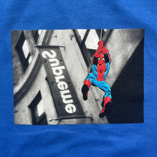 Supreme x Spider-Man Black Tee from the SS26 season is a royal (navy) 100% cotton T-shirt featuring a large box graphic on the front. The print shows a black and white city photo with the Supreme store, while Spider-Man in his red and blue costume swings across the scene in a cartoon-style illustration. The shirt includes a Supreme x Marvel collaboration neck tag.&nbsp;