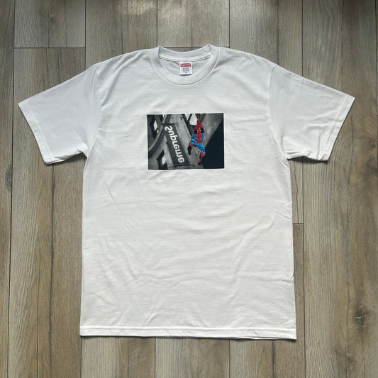 Supreme x Spider-Man Black Tee from the SS26 season is a white 100% cotton T-shirt featuring a large box graphic on the front. The print shows a black and white city photo with the Supreme store, while Spider-Man in his red and blue costume swings across the scene in a cartoon-style illustration. The shirt includes a Supreme x Marvel collaboration neck tag.&nbsp;