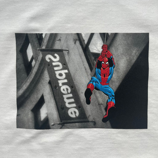 Supreme x Spider-Man Black Tee from the SS26 season is a white 100% cotton T-shirt featuring a large box graphic on the front. The print shows a black and white city photo with the Supreme store, while Spider-Man in his red and blue costume swings across the scene in a cartoon-style illustration. The shirt includes a Supreme x Marvel collaboration neck tag.&nbsp;