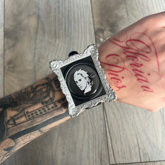 Swatch x Jeremy Scott 2011 Portrait Analog Watch SUOZ121 is an analog watch from the designer’s second collaboration with Swatch, featuring a portrait of Jeremy Scott on the dial and his signature on the strap. The watch includes a silver-tone ornate removable frame, black dial with fixed bezel, and Swiss-made quartz movement. It is water resistant up to 30 meters, comes with original packaging, and retains protective film, with the hour hand partially obscured by the portrait design.
