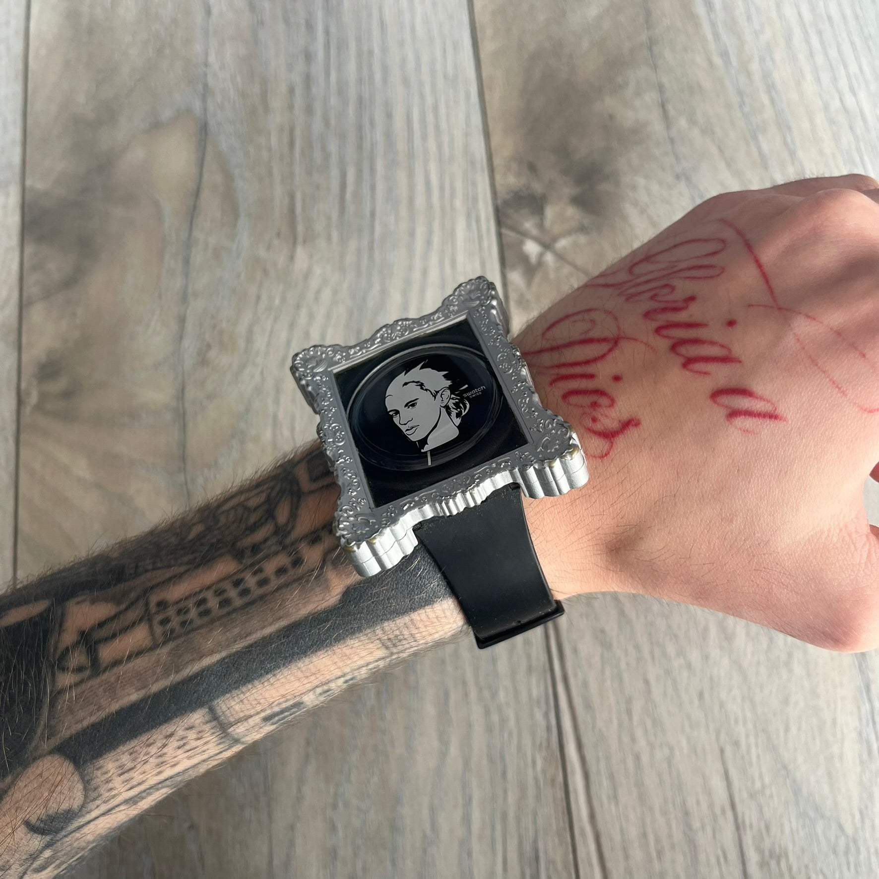 Swatch x Jeremy Scott 2011 Portrait Analog Watch SUOZ121 is an analog watch from the designer’s second collaboration with Swatch, featuring a portrait of Jeremy Scott on the dial and his signature on the strap. The watch includes a silver-tone ornate removable frame, black dial with fixed bezel, and Swiss-made quartz movement. It is water resistant up to 30 meters, comes with original packaging, and retains protective film, with the hour hand partially obscured by the portrait design.