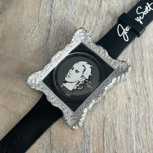 Swatch x Jeremy Scott 2011 Portrait Analog Watch SUOZ121 is an analog watch from the designer’s second collaboration with Swatch, featuring a portrait of Jeremy Scott on the dial and his signature on the strap. The watch includes a silver-tone ornate removable frame, black dial with fixed bezel, and Swiss-made quartz movement. It is water resistant up to 30 meters, comes with original packaging, and retains protective film, with the hour hand partially obscured by the portrait design.