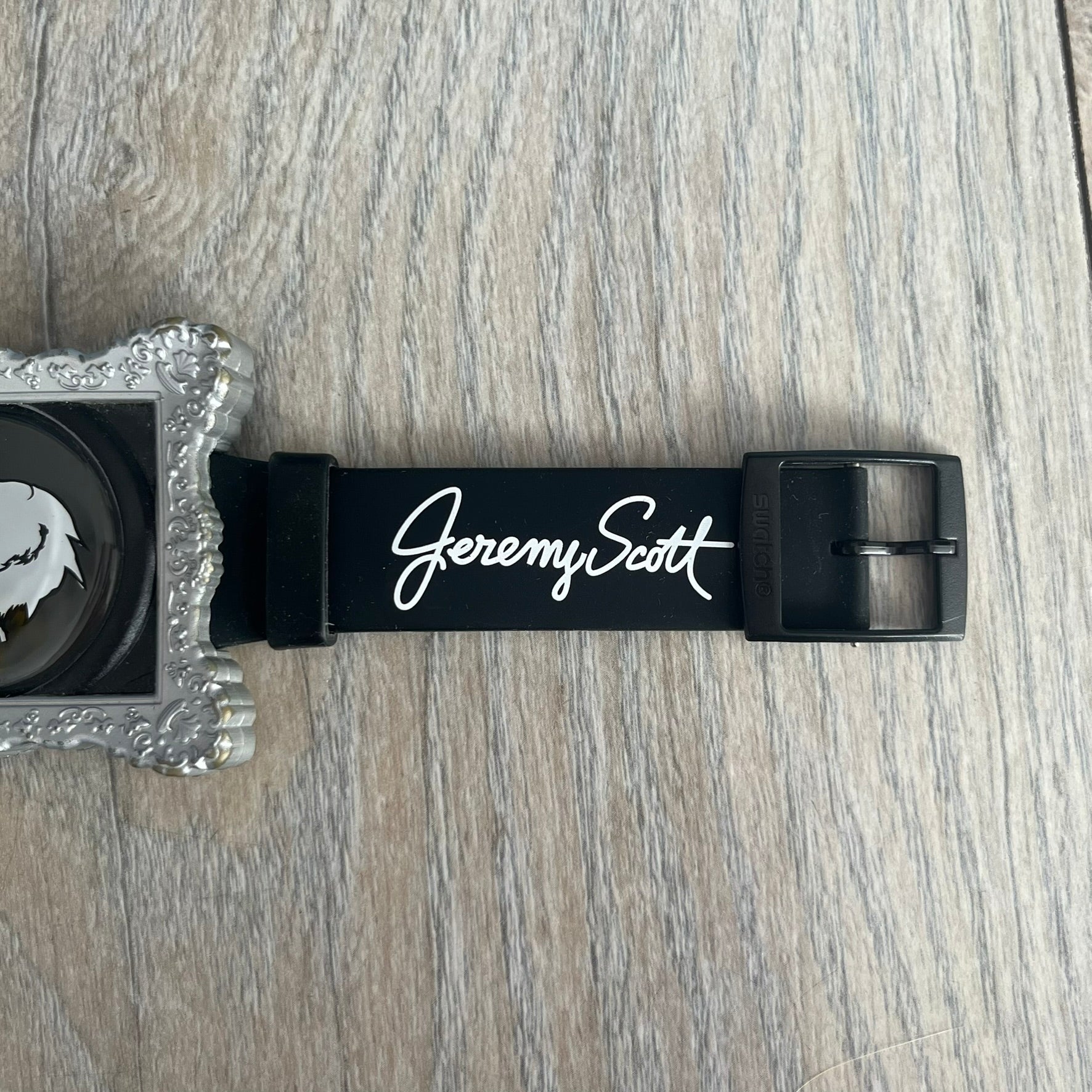 Swatch x Jeremy Scott 2011 Portrait Analog Watch SUOZ121 is an analog watch from the designer’s second collaboration with Swatch, featuring a portrait of Jeremy Scott on the dial and his signature on the strap. The watch includes a silver-tone ornate removable frame, black dial with fixed bezel, and Swiss-made quartz movement. It is water resistant up to 30 meters, comes with original packaging, and retains protective film, with the hour hand partially obscured by the portrait design.