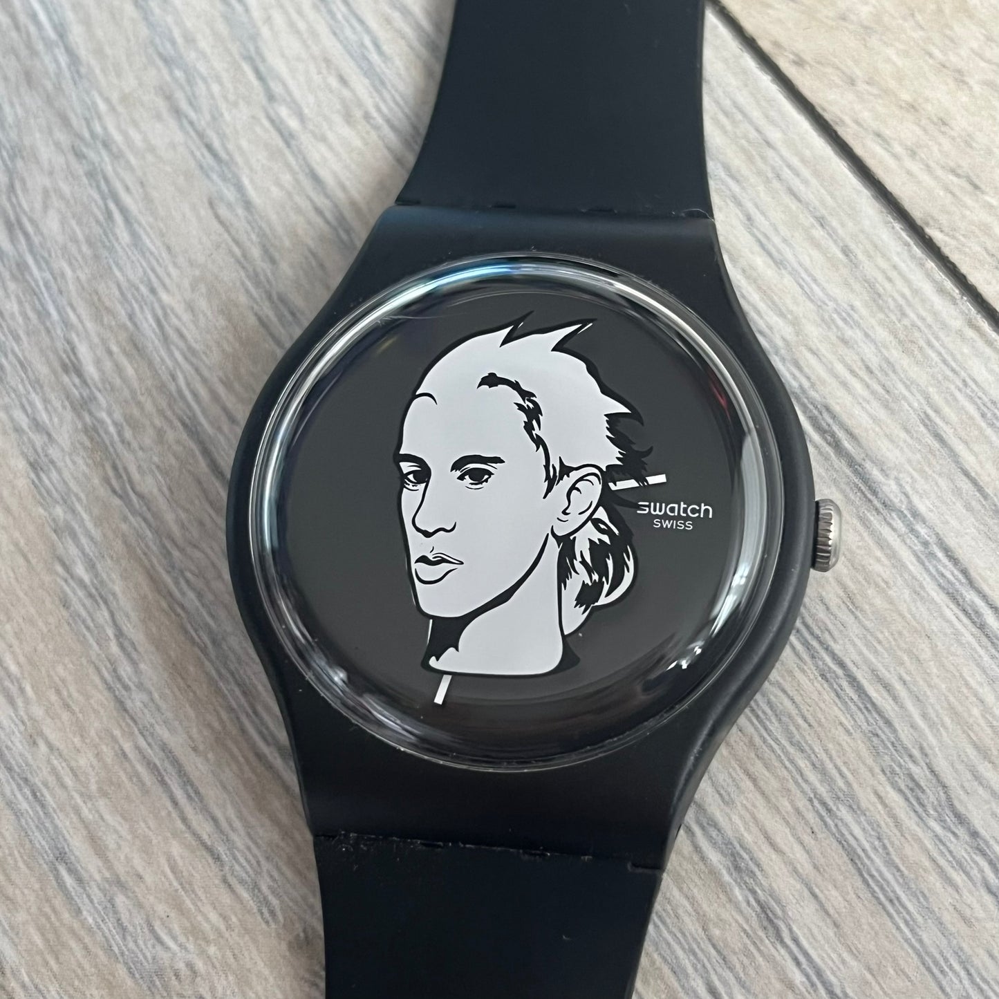 Swatch x Jeremy Scott 2011 Portrait Analog Watch SUOZ121 is an analog watch from the designer’s second collaboration with Swatch, featuring a portrait of Jeremy Scott on the dial and his signature on the strap. The watch includes a silver-tone ornate removable frame, black dial with fixed bezel, and Swiss-made quartz movement. It is water resistant up to 30 meters, comes with original packaging, and retains protective film, with the hour hand partially obscured by the portrait design.
