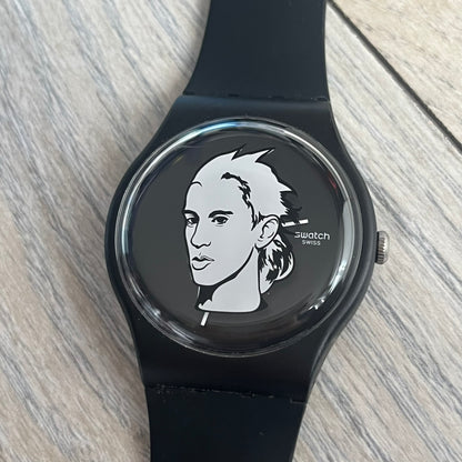 Swatch x Jeremy Scott 2011 Portrait Analog Watch SUOZ121 is an analog watch from the designer’s second collaboration with Swatch, featuring a portrait of Jeremy Scott on the dial and his signature on the strap. The watch includes a silver-tone ornate removable frame, black dial with fixed bezel, and Swiss-made quartz movement. It is water resistant up to 30 meters, comes with original packaging, and retains protective film, with the hour hand partially obscured by the portrait design.