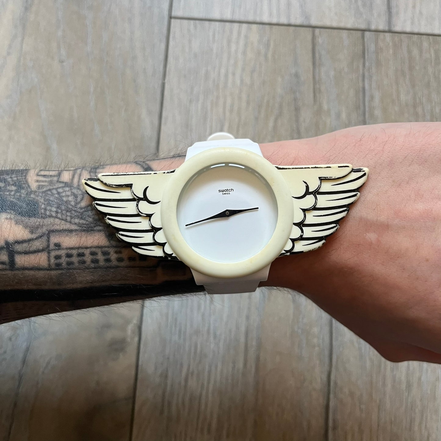 Swatch x Jeremy Scott SUOZ103 “Wings” analog watch released in 2011 as part of Jeremy Scott’s collaboration with Swatch. The watch features a 41 mm case, white dial with Swatch text, black hands, and a white silicone strap. The design includes removable clip-on white 3D wings with black detailing, Swiss quartz movement, patented Swatch battery system, water resistance, and Jeremy Scott branding on the caseback.