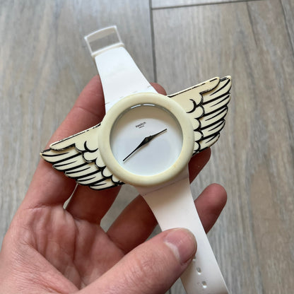 Swatch x Jeremy Scott SUOZ103 “Wings” analog watch released in 2011 as part of Jeremy Scott’s collaboration with Swatch. The watch features a 41 mm case, white dial with Swatch text, black hands, and a white silicone strap. The design includes removable clip-on white 3D wings with black detailing, Swiss quartz movement, patented Swatch battery system, water resistance, and Jeremy Scott branding on the caseback.