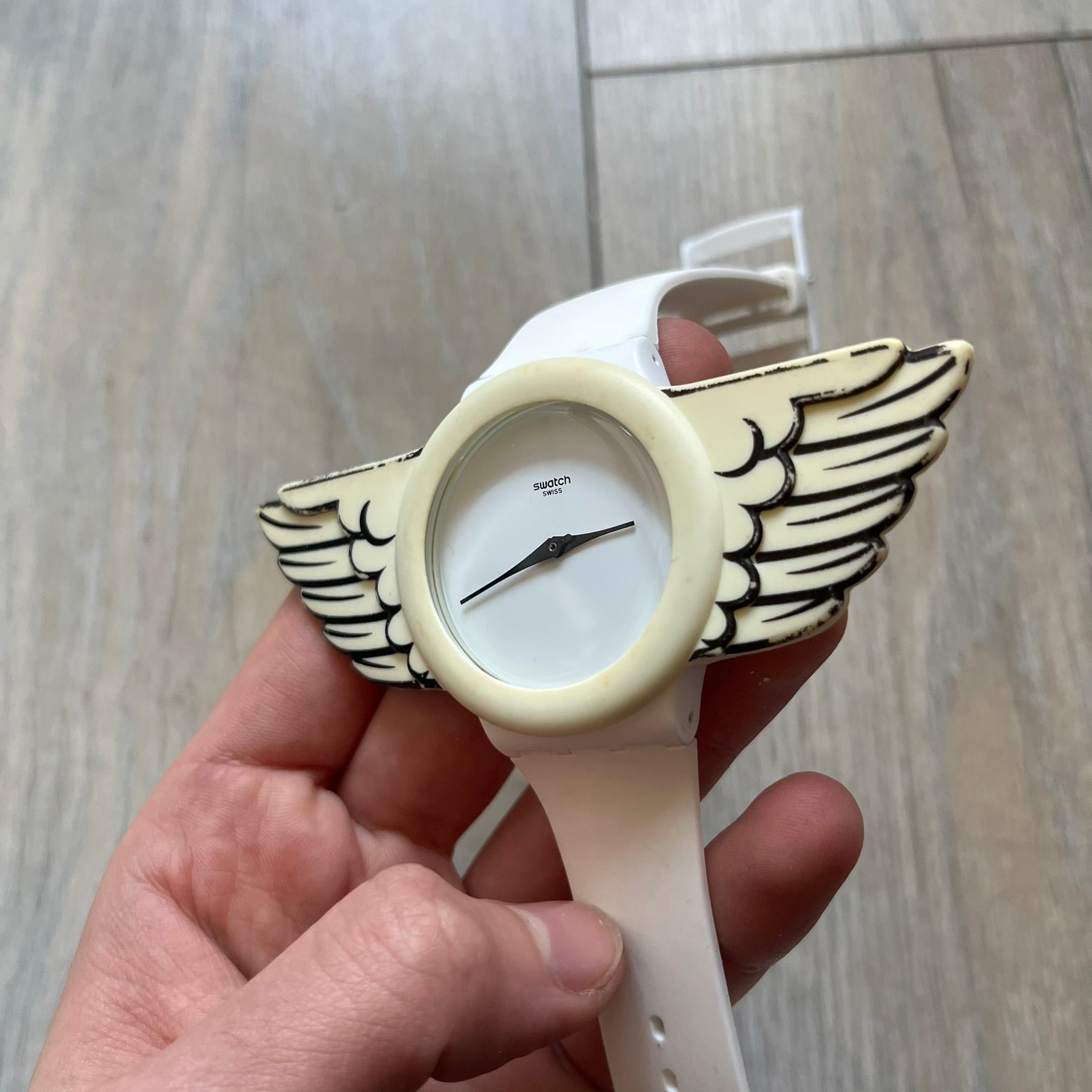 Swatch x Jeremy Scott SUOZ103 “Wings” analog watch released in 2011 as part of Jeremy Scott’s collaboration with Swatch. The watch features a 41 mm case, white dial with Swatch text, black hands, and a white silicone strap. The design includes removable clip-on white 3D wings with black detailing, Swiss quartz movement, patented Swatch battery system, water resistance, and Jeremy Scott branding on the caseback.