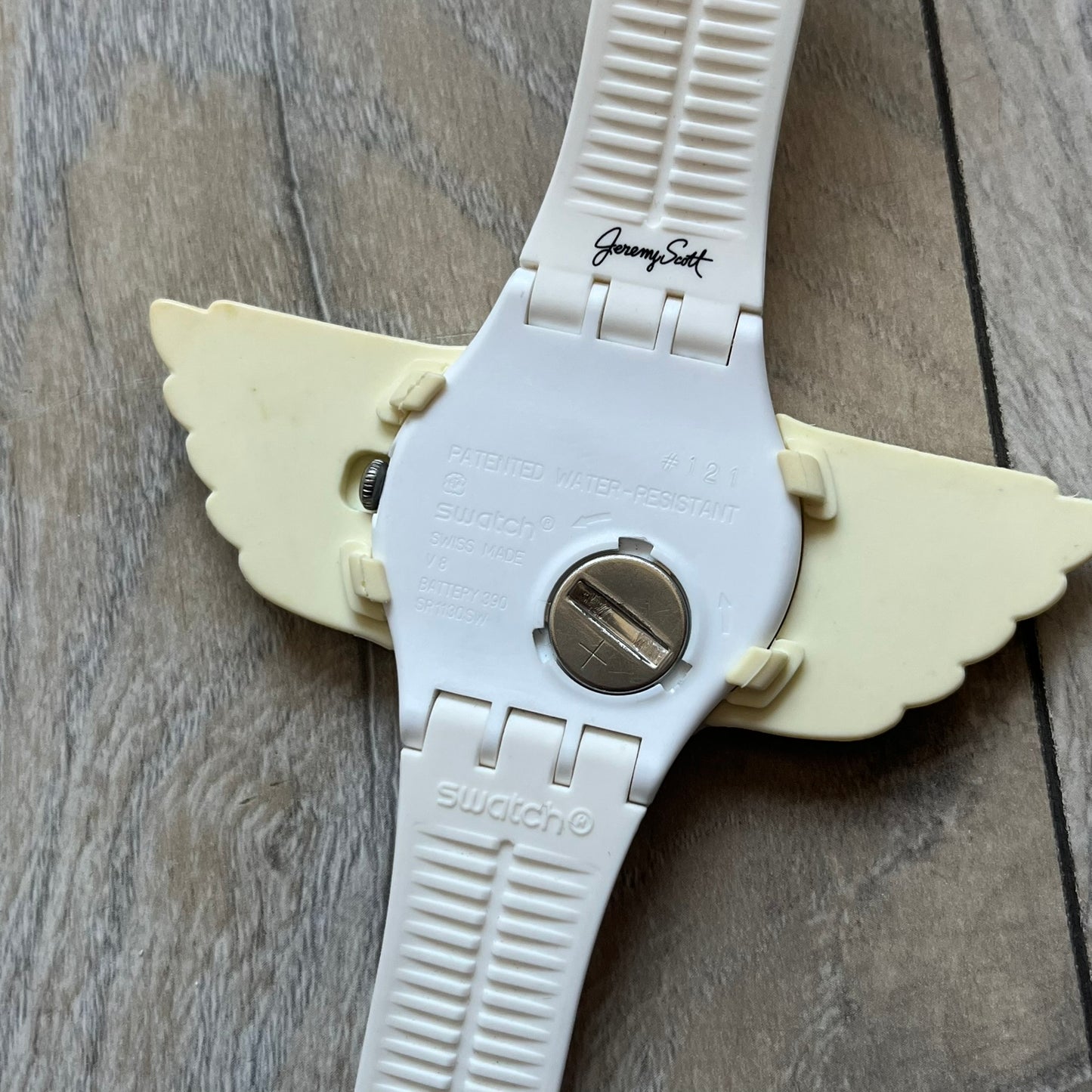Swatch x Jeremy Scott SUOZ103 “Wings” analog watch released in 2011 as part of Jeremy Scott’s collaboration with Swatch. The watch features a 41 mm case, white dial with Swatch text, black hands, and a white silicone strap. The design includes removable clip-on white 3D wings with black detailing, Swiss quartz movement, patented Swatch battery system, water resistance, and Jeremy Scott branding on the caseback.