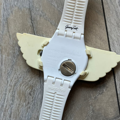 Swatch x Jeremy Scott SUOZ103 “Wings” analog watch released in 2011 as part of Jeremy Scott’s collaboration with Swatch. The watch features a 41 mm case, white dial with Swatch text, black hands, and a white silicone strap. The design includes removable clip-on white 3D wings with black detailing, Swiss quartz movement, patented Swatch battery system, water resistance, and Jeremy Scott branding on the caseback.