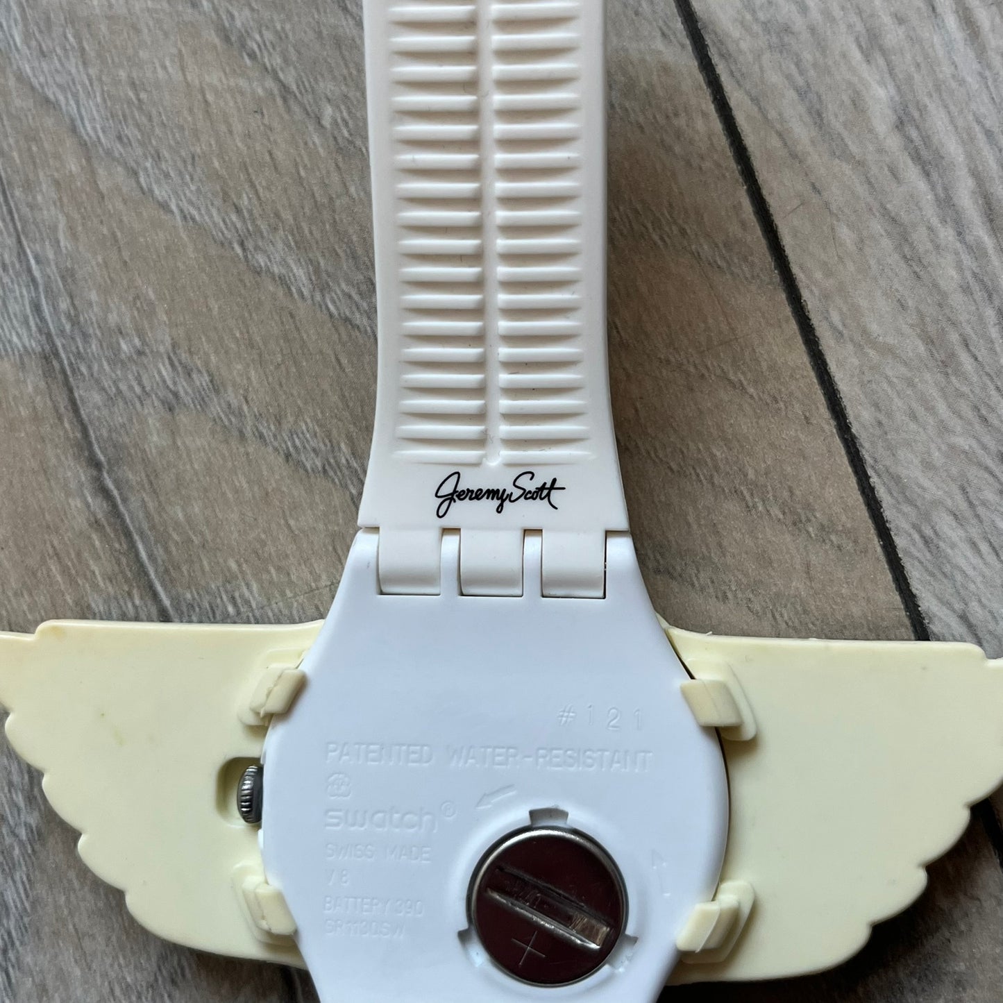 Swatch x Jeremy Scott SUOZ103 “Wings” analog watch released in 2011 as part of Jeremy Scott’s collaboration with Swatch. The watch features a 41 mm case, white dial with Swatch text, black hands, and a white silicone strap. The design includes removable clip-on white 3D wings with black detailing, Swiss quartz movement, patented Swatch battery system, water resistance, and Jeremy Scott branding on the caseback.