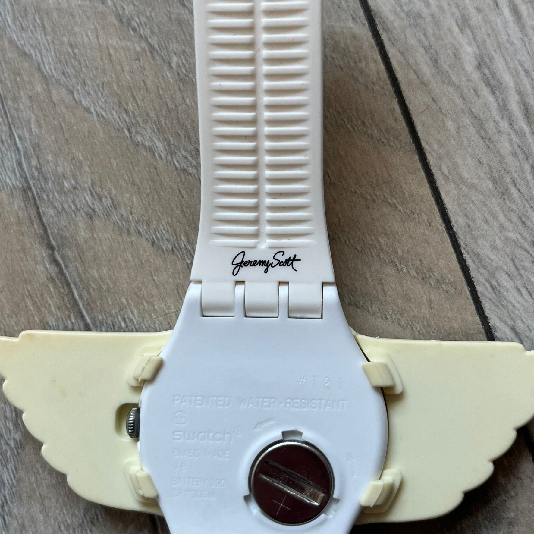 Swatch x Jeremy Scott SUOZ103 “Wings” analog watch released in 2011 as part of Jeremy Scott’s collaboration with Swatch. The watch features a 41 mm case, white dial with Swatch text, black hands, and a white silicone strap. The design includes removable clip-on white 3D wings with black detailing, Swiss quartz movement, patented Swatch battery system, water resistance, and Jeremy Scott branding on the caseback.