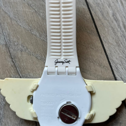 Swatch x Jeremy Scott SUOZ103 “Wings” analog watch released in 2011 as part of Jeremy Scott’s collaboration with Swatch. The watch features a 41 mm case, white dial with Swatch text, black hands, and a white silicone strap. The design includes removable clip-on white 3D wings with black detailing, Swiss quartz movement, patented Swatch battery system, water resistance, and Jeremy Scott branding on the caseback.
