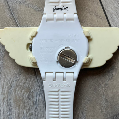 Swatch x Jeremy Scott SUOZ103 “Wings” analog watch released in 2011 as part of Jeremy Scott’s collaboration with Swatch. The watch features a 41 mm case, white dial with Swatch text, black hands, and a white silicone strap. The design includes removable clip-on white 3D wings with black detailing, Swiss quartz movement, patented Swatch battery system, water resistance, and Jeremy Scott branding on the caseback.