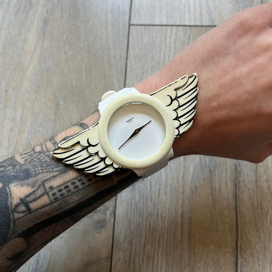 Swatch x Jeremy Scott SUOZ103 “Wings” analog watch released in 2011 as part of Jeremy Scott’s collaboration with Swatch. The watch features a 41 mm case, white dial with Swatch text, black hands, and a white silicone strap. The design includes removable clip-on white 3D wings with black detailing, Swiss quartz movement, patented Swatch battery system, water resistance, and Jeremy Scott branding on the caseback.