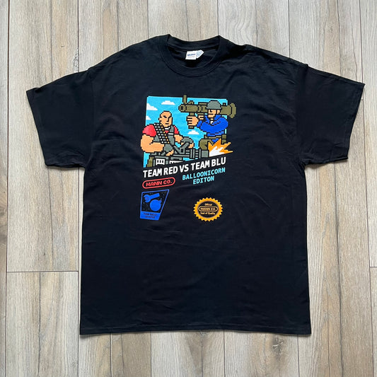 Team Fortress 2 Arcade 2014 Nerdblock Tee is a black cotton T-shirt released as part of a Nerdblock promotional collaboration with Valve. The front features an arcade-style pixel graphic of Red Team Heavy and Blue Team Soldier in combat. Below the artwork is “Team Red vs Team Blu” text along with Mann Co branding, including a red logo, “Blue Balloonicorn Edition” text, and a “Team Based Shooter” label. The design is completed with a yellow official Mann Co. Seal of Quality and a Gildan blank base.
