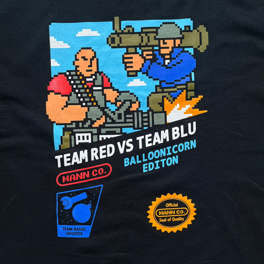 Team Fortress 2 Arcade 2014 Nerdblock Tee is a black cotton T-shirt released as part of a Nerdblock promotional collaboration with Valve. The front features an arcade-style pixel graphic of Red Team Heavy and Blue Team Soldier in combat. Below the artwork is “Team Red vs Team Blu” text along with Mann Co branding, including a red logo, “Blue Balloonicorn Edition” text, and a “Team Based Shooter” label. The design is completed with a yellow official Mann Co. Seal of Quality and a Gildan blank base.
