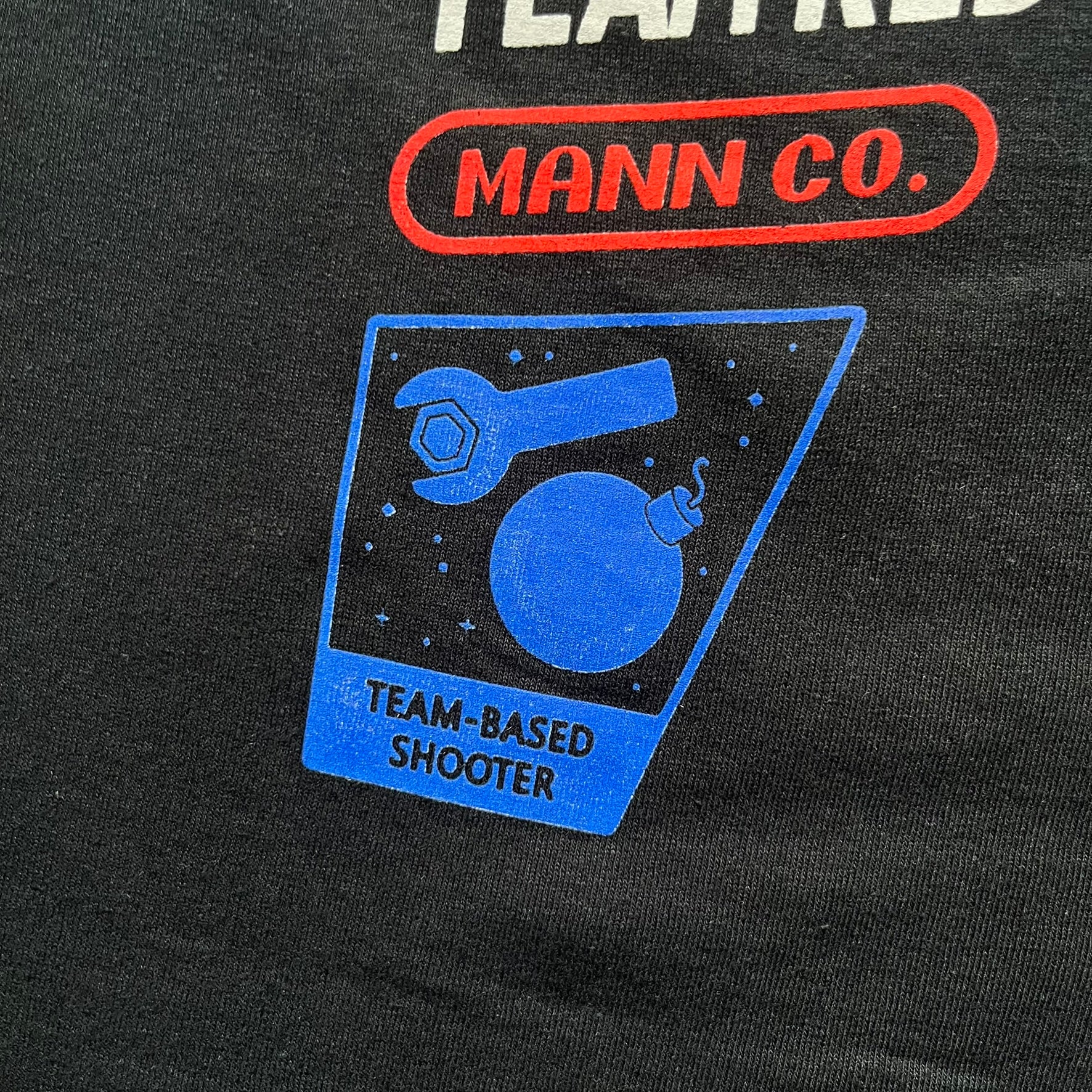 Team Fortress 2 Arcade 2014 Nerdblock Tee is a black cotton T-shirt released as part of a Nerdblock promotional collaboration with Valve. The front features an arcade-style pixel graphic of Red Team Heavy and Blue Team Soldier in combat. Below the artwork is “Team Red vs Team Blu” text along with Mann Co branding, including a red logo, “Blue Balloonicorn Edition” text, and a “Team Based Shooter” label. The design is completed with a yellow official Mann Co. Seal of Quality and a Gildan blank base.
