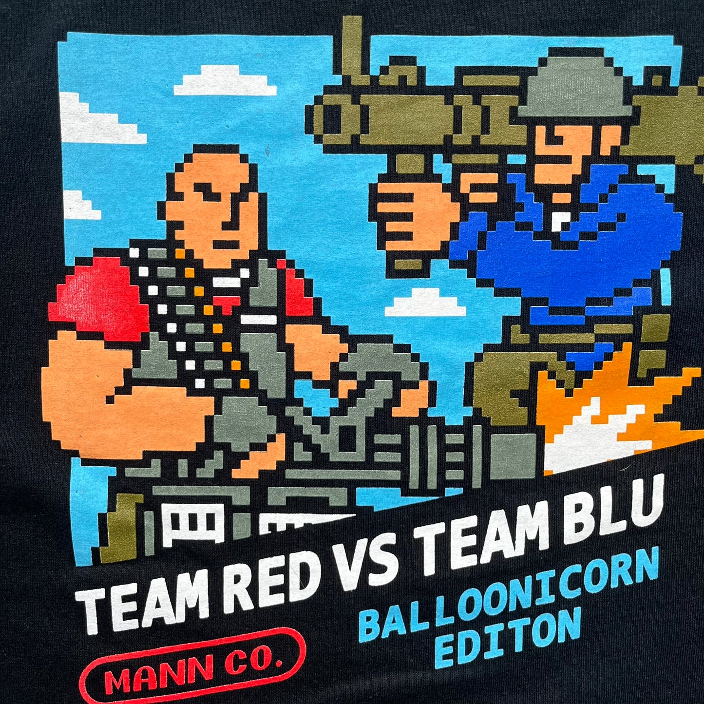 Team Fortress 2 Arcade 2014 Nerdblock Tee is a black cotton T-shirt released as part of a Nerdblock promotional collaboration with Valve. The front features an arcade-style pixel graphic of Red Team Heavy and Blue Team Soldier in combat. Below the artwork is “Team Red vs Team Blu” text along with Mann Co branding, including a red logo, “Blue Balloonicorn Edition” text, and a “Team Based Shooter” label. The design is completed with a yellow official Mann Co. Seal of Quality and a Gildan blank base.
