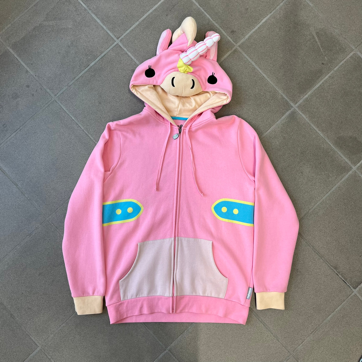 Team Fortress 2 Balloonicorn 2015 WeLoveFine Zip-Up Hoodie is a bright pink unisex colorblock hoodie from Valve’s Team Fortress 2 line, with a detailed 3D plush Balloonicorn hood featuring embroidered eyes, a 3D horn, nose, and ears. It has front pockets, a custom metal Balloonicorn cameo zipper pull, and a light blue printed saddle graphic that wraps from the back to the front. The licensed TF2 neck tag carries 2015 and We Love Fine branding. Super rare.