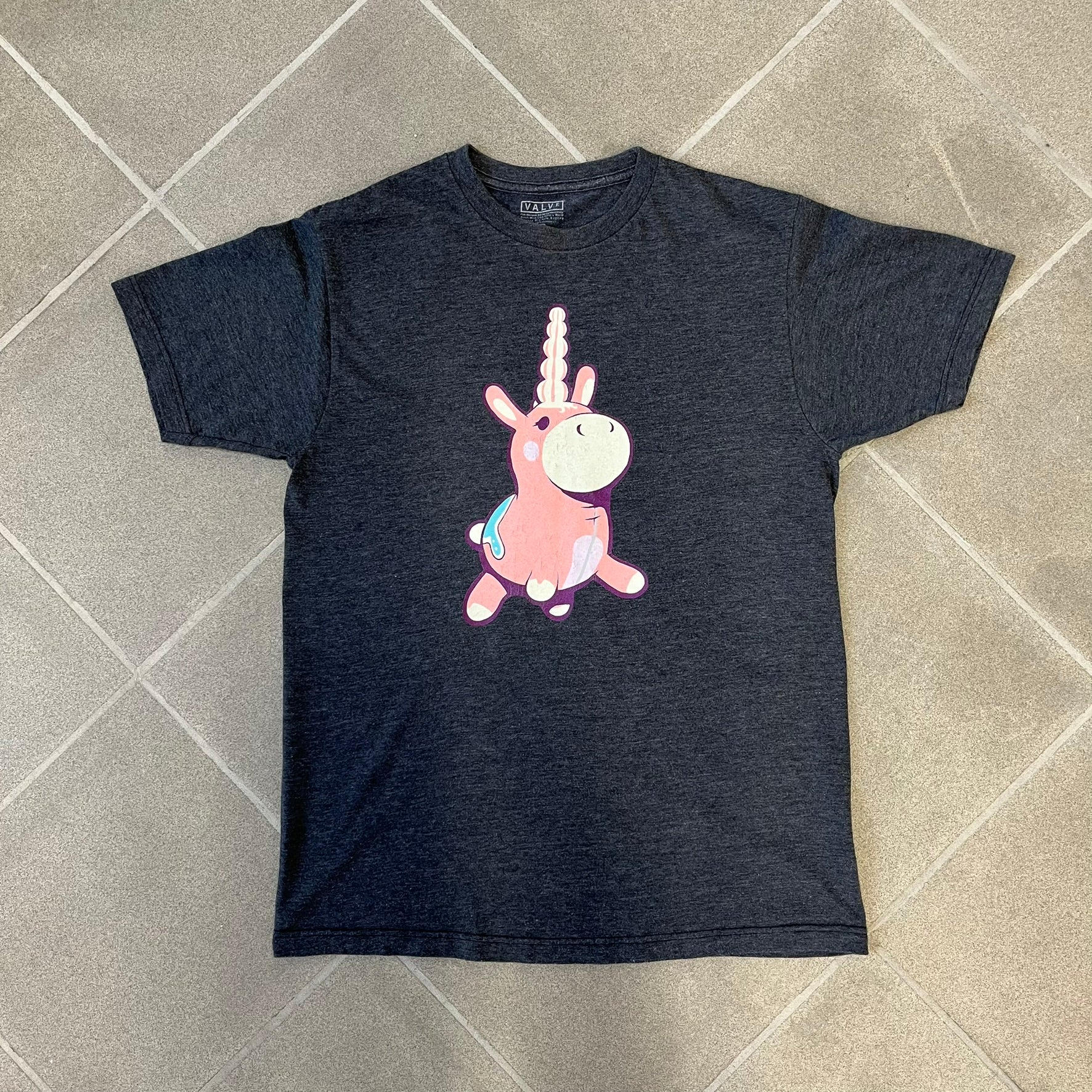 Team Fortress 2 “Balloonicorn & Sentry” 2010s Tee is a dark grey cotton-polyester blend T-shirt with a printed Valve neck tag. The front features a pink Balloonicorn graphic, while the back displays a red Engineer Sentry graphic. This officially licensed Team Fortress 2 T-shirt is a rare release.
